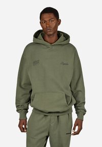 Oliwkowa bluza z kapturem, z przednią kieszenią, ściągaczami na rękawach i u dołu. Posiada dwa haftowane logo: "Whispers" oraz "Pegador". Miękka w dotyku.