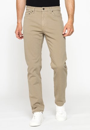 Uomo con pantaloni beige casual e scarpe da ginnastica bianche, in piedi su sfondo bianco.