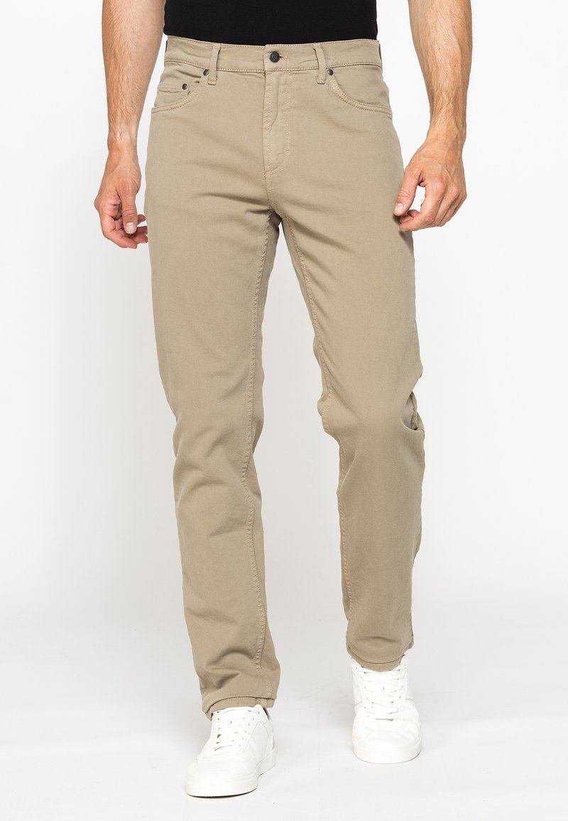 Uomo con pantaloni beige casual e scarpe da ginnastica bianche, in piedi su sfondo bianco.
