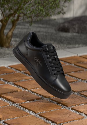 Svarta läder sneakers från Calvin Klein med snören, svävande över en golv av trämosaik och små stenar, med ett träd och en vägg i bakgrunden.