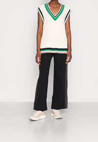 Haut blanc sans manches à côtes avec des rayures vertes, bleu marine et rouges ; associé à un pantalon large noir avec des rayures latérales et des baskets claires.