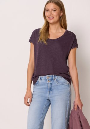 Kvinde smilende, iført en lilla kortærmet top og lyseblå jeans, holder en sammenfoldet lilla sweater i venstre hånd.