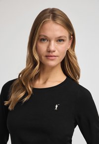 Svart långärmad tröja med en liten broderad golfspelardesign i vitt. Mjukt, figursytt material med rund halsringning.