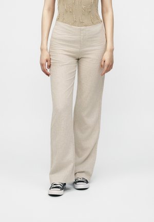 Person iført beige bukser med vide ben, en lysebrun, tekstureret ærmeløs top og sorte canvas-sneakers står mod en ensfarvet baggrund.