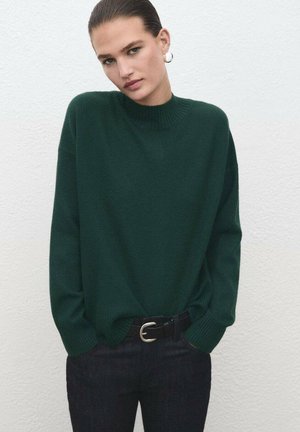 Jersey de punto verde oscuro con cuello alto, mangas largas y puños de canalé, combinado con unos vaqueros oscuros y un cinturón con una hebilla plateada.