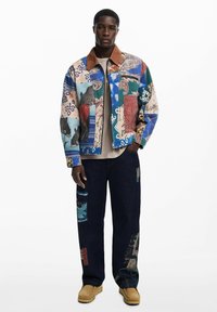 Kirjus denim jakk, millel on mitmevärvilised kangamustrid, pruuni korduroo kraega, koos tume-denimist pükstega, mis on kaunistatud kangapatchidega.