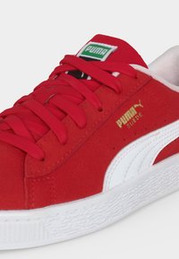 Κόκκινο sneaker Puma από σουέτ με λευκή σόλα και ρίγα, χρυσό λογότυπο Puma στο πλάι και κόκκινα κορδόνια, εμφανίζεται από γωνιακή κοντινή λήψη.