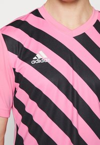 Maglietta sportiva a righe rosa e nere con scollo a V, realizzata in tessuto liscio. Presenta un piccolo logo Adidas bianco sul petto a sinistra.