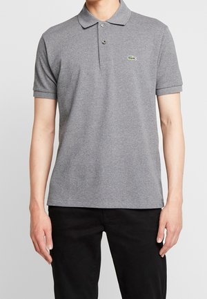 Homme portant un polo gris à manches courtes avec col et boutons, assorti à un pantalon noir, debout devant un fond uni.