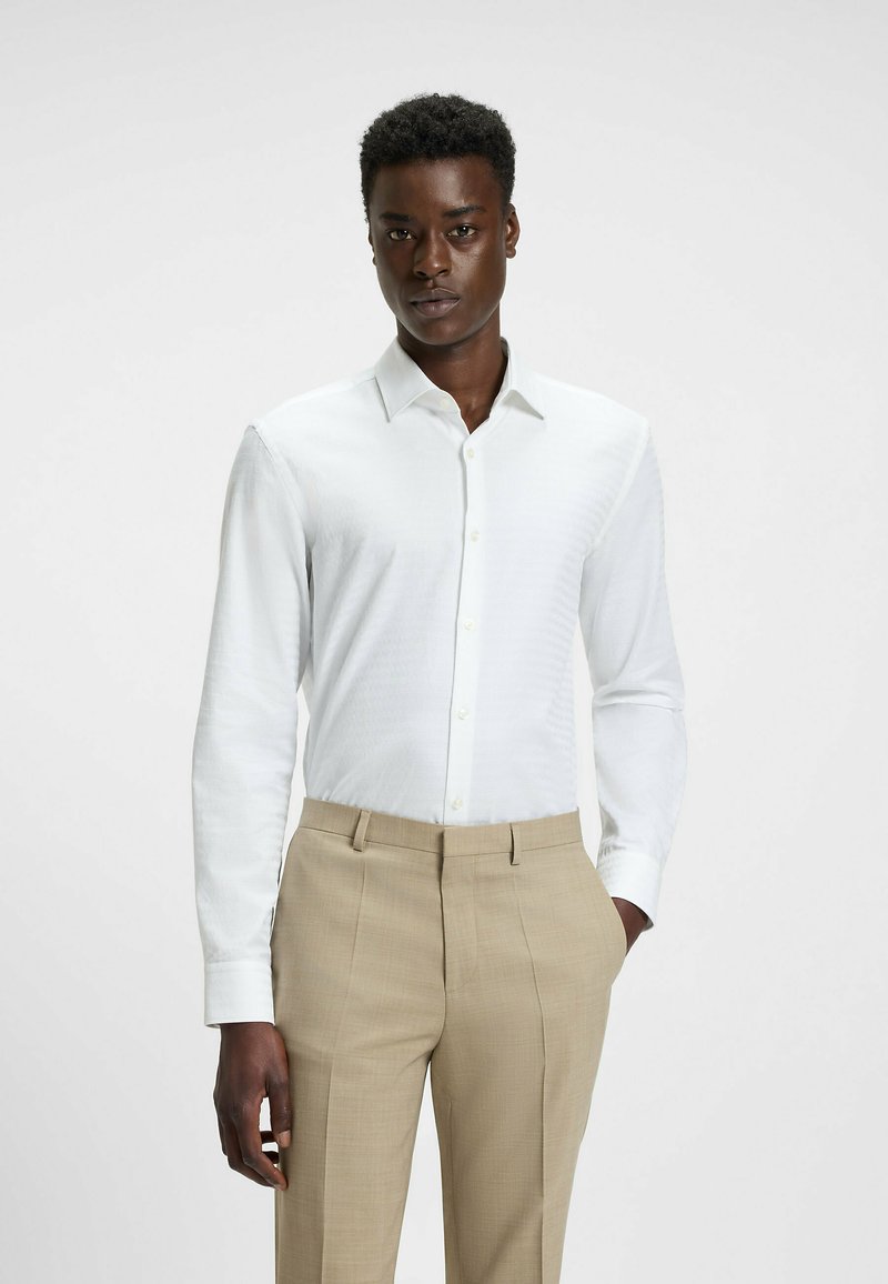 Witte blouse met lange mouwen en een subtiel ruitpatroon, gecombineerd met beige pantalon. De blouse heeft een klassieke kraag en een knopenfront.