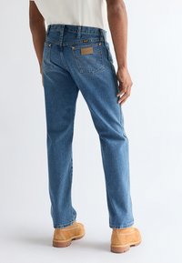 Person med blå jeans med rette buksebein og brune arbeidssko, stående med ryggen vendt mot kameraet foran en ensfarget hvit bakgrunn.