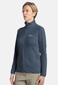 Veste zippée bleu foncé en tissu texturé avec un col haut, dotée d'un logo sur la poitrine gauche et de poches latérales.