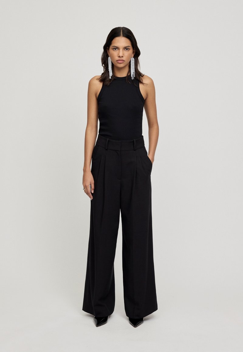 Top preto sem mangas combinado com calças pretas de cintura alta e perna larga. Brincos compridos com contas, sapatos pretos elegantes de bico fino.