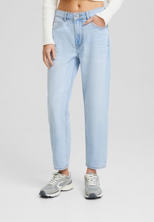 Persoon draagt lichtblauwe high-waist rechte jeans met voorzakken, witte cropped trui en grijs-witte sneakers staand.