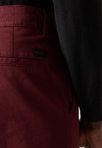 Pantalons bordeaux avec un léger éclat, présentant une poche arrière et une étiquette noire avec "BOSS" en lettres dorées. Gros plan sur la texture du tissu.