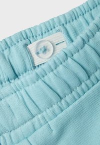 Jasnoniebieska tkanina w pasie z gumką i widocznymi szwami; zawiera przezroczysty guzik oraz tkaną metkę z dziurką na guzik.