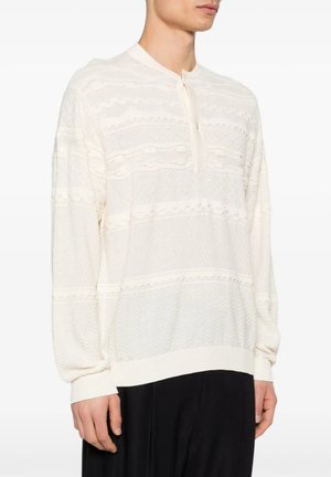 Uomo che indossa un maglione bianco lavorato a maglia a maniche lunghe con una corta patta con bottoni, abbinato a pantaloni neri, mostrato dal collo ai fianchi.