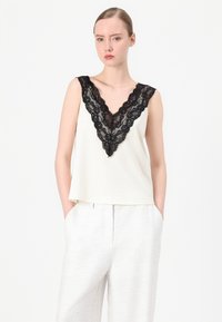 Cream-colored sleeveless top with a black lace V-neckline, featuring a relaxed fit and smooth texture, paired with light, textured trousers.