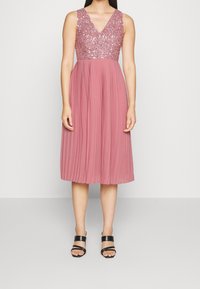 Sleeveless pink dress with a V-neck, embellished sequin bodice, and pleated skirt. Worn with black strappy sandals.