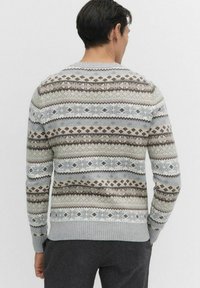 Man met de rug naar de kijker, gekleed in een lichtgrijze trui met horizontale Noorse patronen in bruin, beige en wit, gecombineerd met donkergrijze broek.