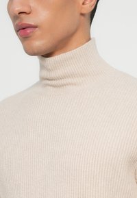 Nahaufnahme einer Person, die einen beigen gerippten Rollkragenpullover trägt. Zu sehen sind der Nacken, die Kieferlinie und die Lippen vor einem weißen Hintergrund.