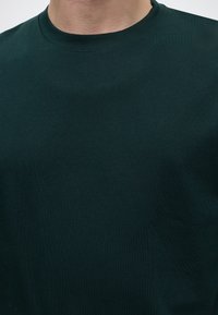 Homme portant un t-shirt vert foncé à col rond, visible des épaules au cou sur un fond uni.