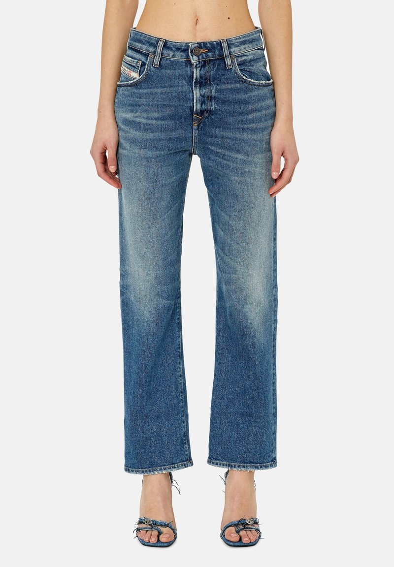 Femme portant un jean bleu taille haute, jambes droites, ourlet effiloché et des sandales en denim à bout ouvert sur fond uni.