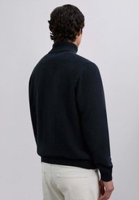 Jersey de cuello alto azul marino con un cuello y puños acanalados, que presenta una textura suave y un corte relajado. Se muestra desde la parte de atrás, combinado con pantalones claros.