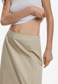 Beige skirt made of smooth fabric with a diagonal seam, featuring a side zipper. Model wears a fitted white ribbed top.