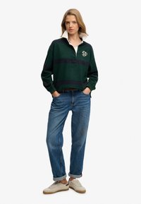 Jeune femme debout, mains dans les poches, portant un polo rayé vert et marine, un jean bleu aux revers retroussés et des baskets beiges sur fond blanc.