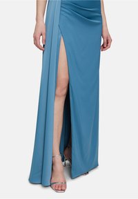 Himmelblaues, bodenlanges Kleid mit einem seitlichen Schlitz und drapiertem Detail. Glatter Stoff mit einem leichten Glanz. Kombiniert mit schimmernden silbernen Sandalen.
