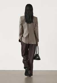 Personne aux longs cheveux noirs porte un blazer beige et un pantalon foncé, marchant en s'éloignant tout en tenant un petit sac à main noir dans sa main droite.