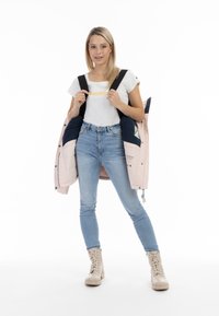 Chaqueta acolchada rosa claro con detalles en azul marino, llevada abierta sobre una camisa blanca. Jeans ajustados de mezclilla azul claro y botas de cordones beige.