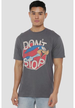 Junger Mann, der ein graues T-Shirt mit einem bunten Cartoon-Vogel und dem Text "Don't Stop," trägt, steht vor einem schlichten hellen Hintergrund.