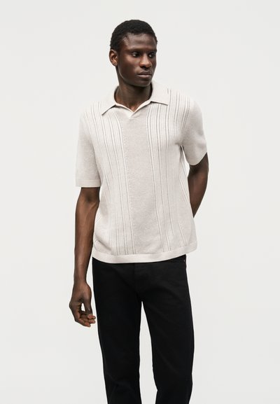Άνδρας φοράει μπεζ πλεκτή πόλο μπλούζα με κοντά μανίκια και κάθετα σχέδια, καθώς και μαύρο παντελόνι, στέκεται μπροστά σε απλό λευκό φόντο.