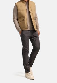 Hombre vestido con un chaleco acolchado beige, suéter marrón claro, pantalones gris oscuro y zapatillas beige, de pie con las manos en los bolsillos sobre un fondo blanco.