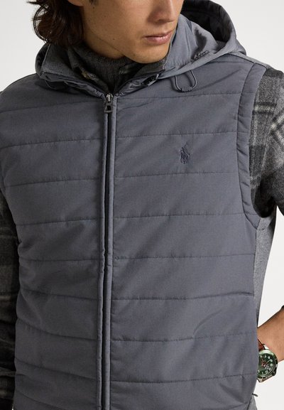 Grå quiltet vest med hætte, udstyret med lynlås og diskret logodetalje. Bruges over en tekstureret sweater, med et synligt ur.