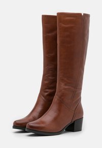 Botas de cuero marrón hasta la rodilla con tacón cuadrado, que presentan una textura suave, puntera redonda, acentos de costura y un pequeño corte en la parte superior.