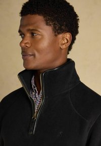 Uomo con capelli ricci corti che indossa un pile nero con mezze zip sopra una camicia a scacchi rossa e bianca, rivolto a sinistra su uno sfondo neutro.