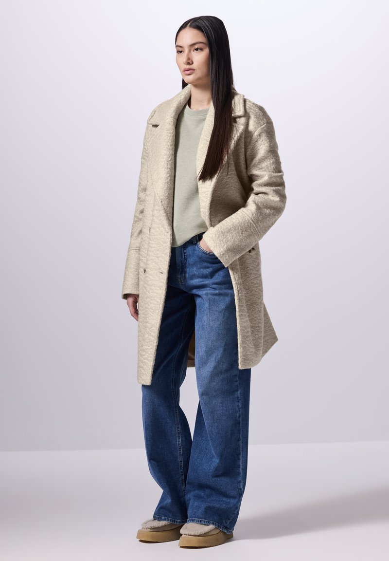 Cappotto in misto lana beige con una finitura testurizzata, indossato sopra un top verde chiaro e jeans a gamba larga blu. Include calzature color beige con una fodera morbida.
