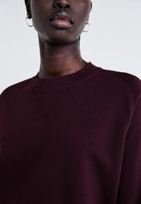 Sweatshirt bordeaux avec col rond côtelé et détails de couture aux épaules, présentant une texture douce et un design minimaliste.