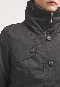 Svart vadderad jacka med hög krage, med en framdragkedja, två knappstängningar och ett slätt, strukturerat tyg.