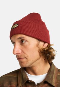Berretto lavorato a maglia rosso con una texture a coste e una parte superiore rotonda. Presenta un'etichetta tessuta circolare e piccola sulla parte anteriore.