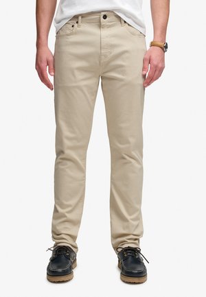 Mann trägt beige Slim-Fit-Hose, weißes Hemd eingesteckt, schwarze Schnürschuhe und eine Armbanduhr mit einem hellbraunen Lederarmband.