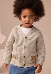 Cardigan en maille crème avec texture côtelée, trois boutons, et appliqué d'ours sur les poches. Le mannequin porte une chemise blanche et un short en jean.