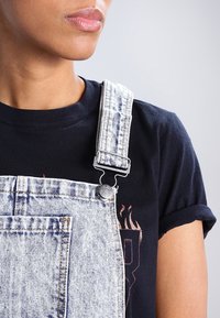 Närbild av en person som bär ljusblå denimoverall över en svart t-shirt med uppvikta ärmar, som visar axelremmen och en del av bröstområdet.