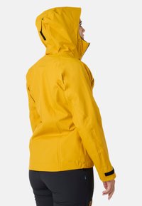 Gul vandafvisende jakke med justerbar hætte, lynlås foran og velcro-stropper ved manchetterne. Har et slankt, tætsiddende design og en glat overflade.