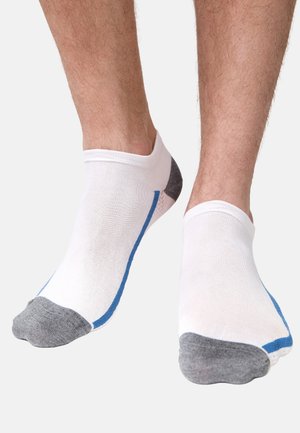 Füße mit weißen Knöchelsocken, die graue Zehen und Fersen sowie einen dünnen blauen Streifen entlang der Seite haben, vor einem einfarbigen hellen Hintergrund.