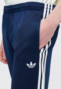 Tengerészkék jogging nadrág elasztikus derékpánttal, oldalzsebekkel és három fehér csíkkal az oldalak mentén. A bal combon fehér logóval.