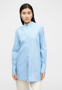 Γυναίκα που φοράει μπλούζα με μακριά μανίκια, κατακόρυφες ρίγες σε μπλε και άσπρο, με μανδύα μανδαρίνου, συνδυασμένη με ανοιχτόχρωμο παντελόνι.